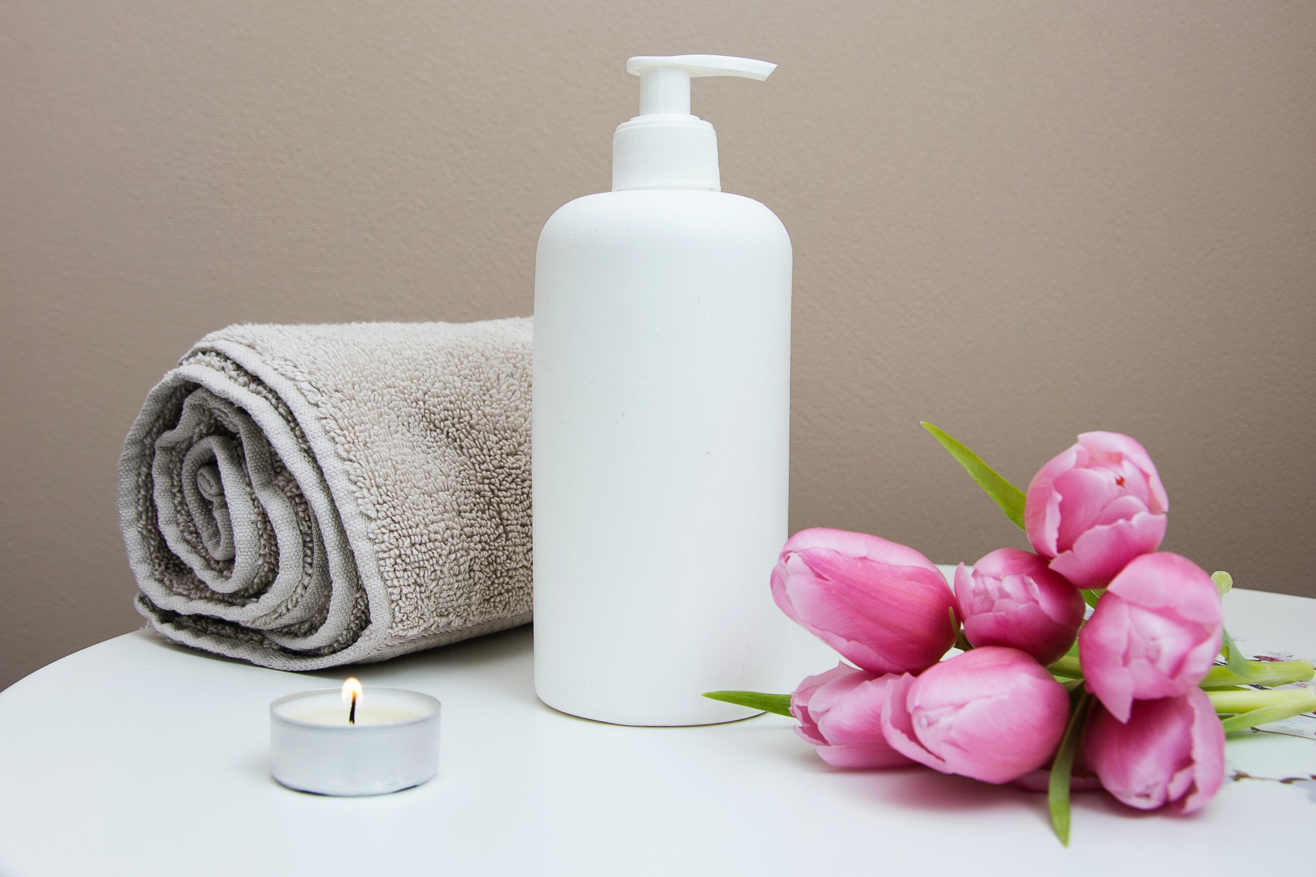 A light brown towel rolled up next to a white lotion bottle. There is a small night light and pink rose on other side of the lotion
