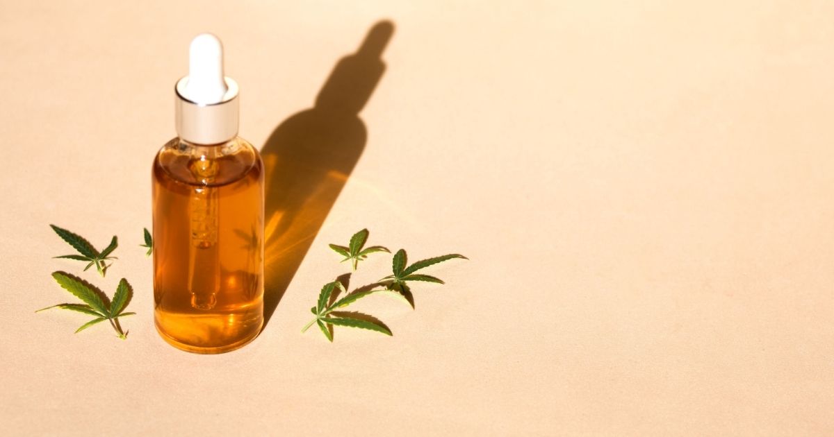 A brown glass bottle with a white lid stands on a pale background, with green leaves on two sides.