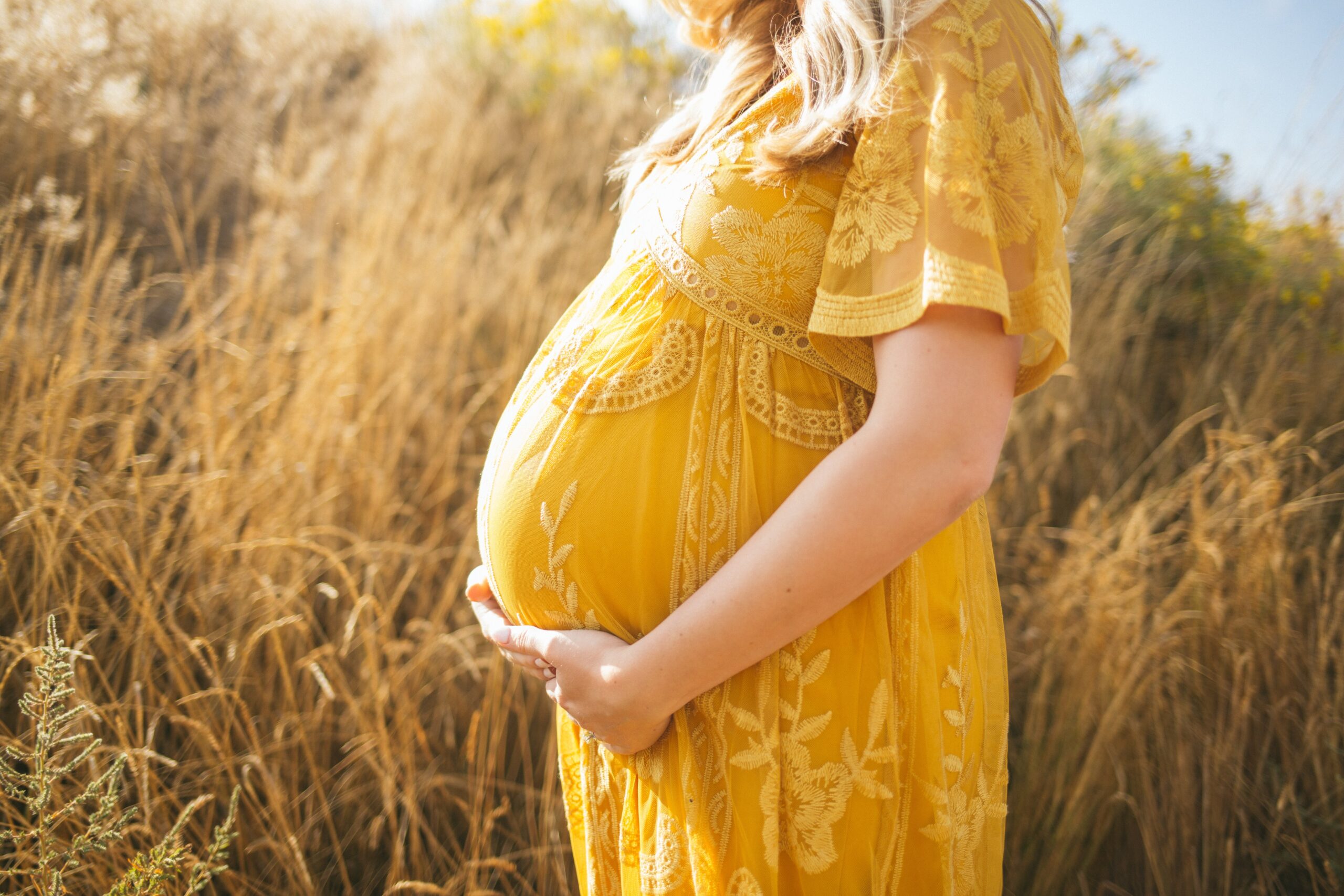 A woman in a yellow dress holding a pregnancy bump while standing in a field