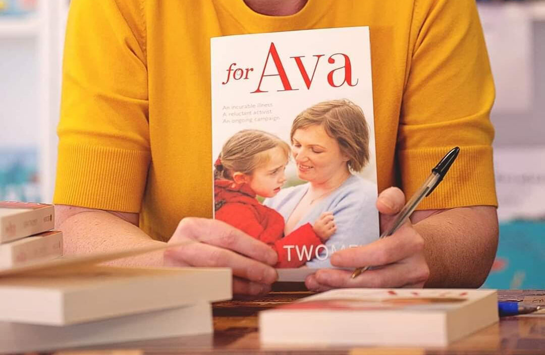 A person wearing a yellow sweater holds the cover of a book to the camera