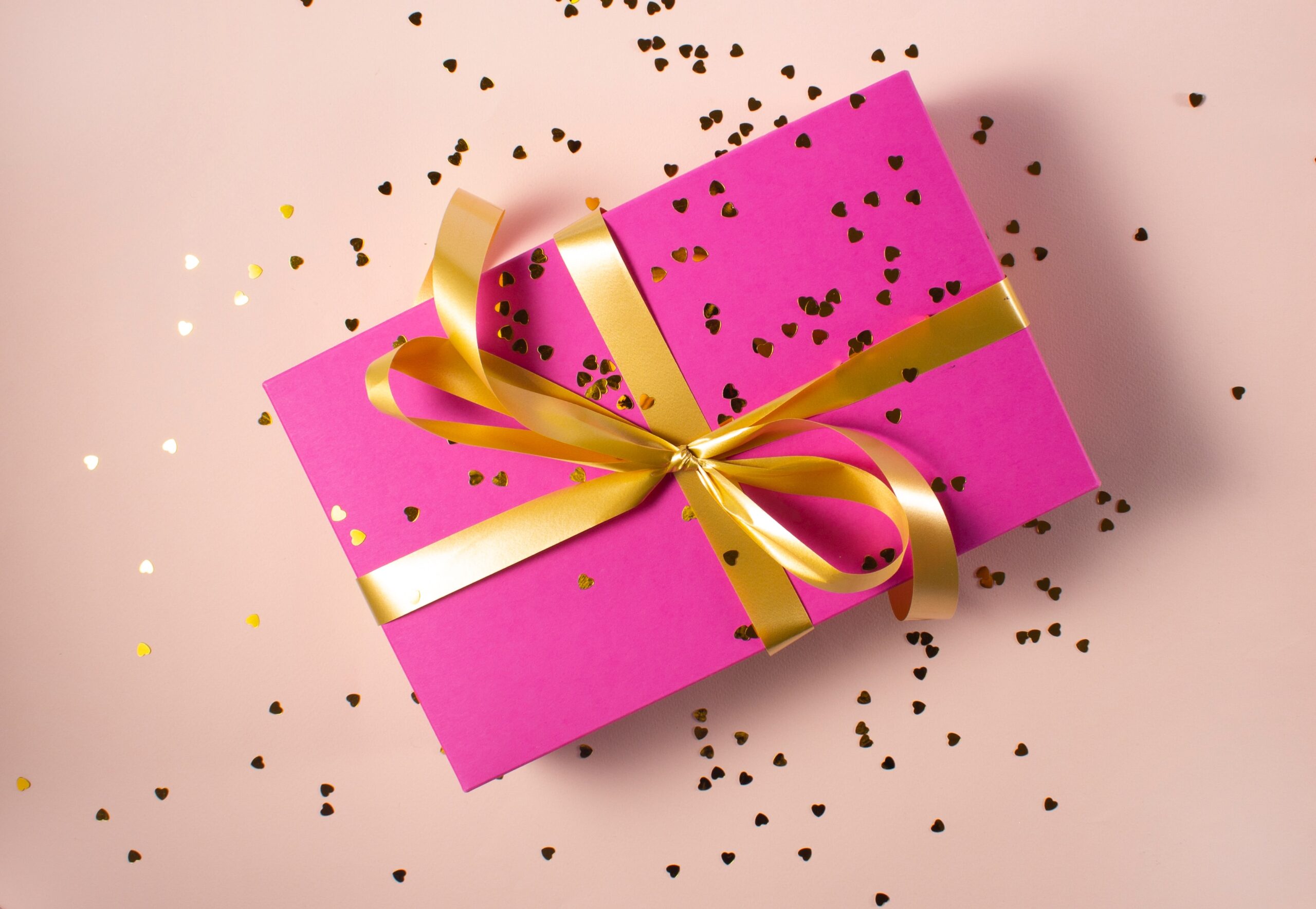 A present wrapped in pink paper with a gold ribbon and bow. There is glitter sprinkled across the top