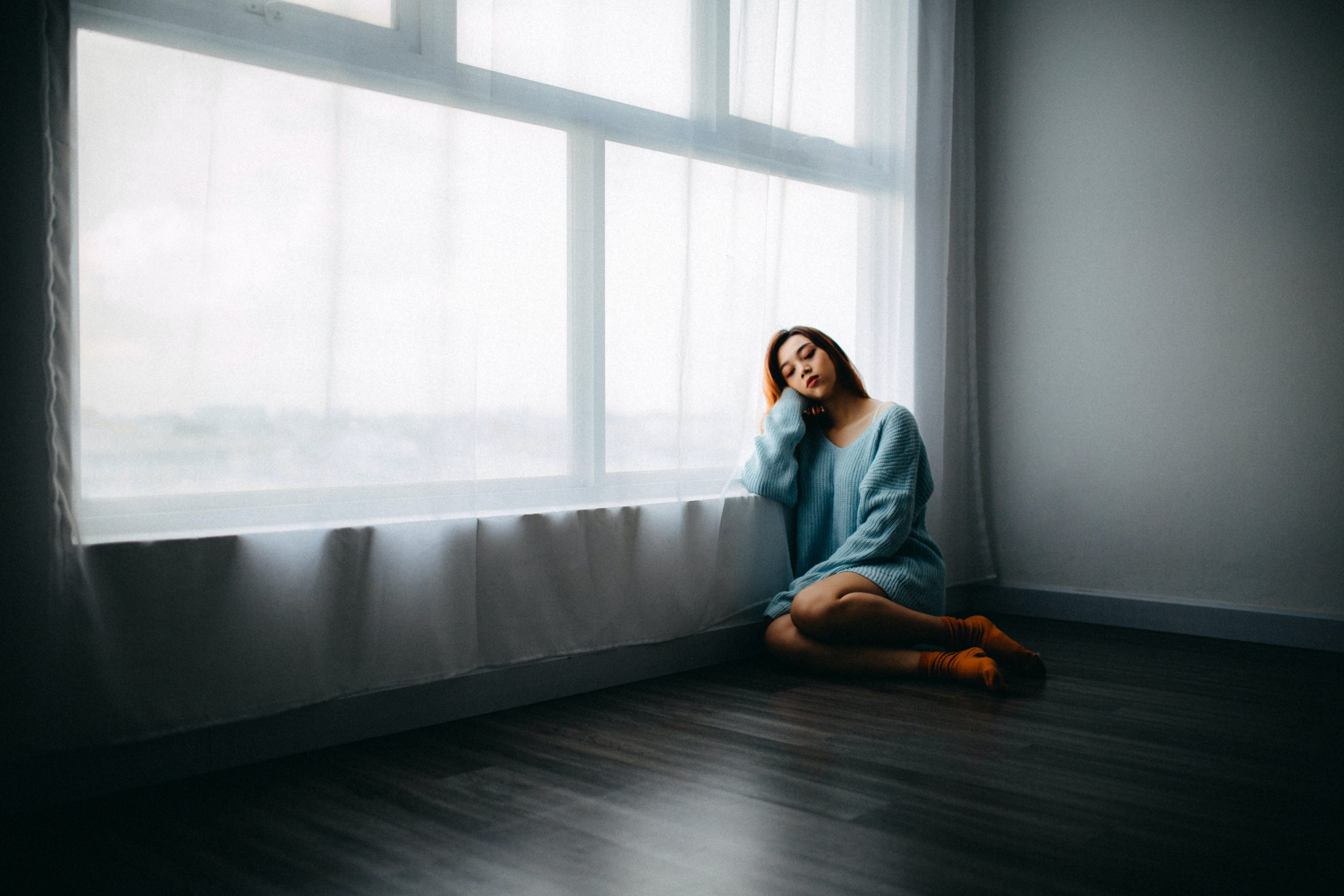 A woman sits on the floor near a large window covered with a voile curtain