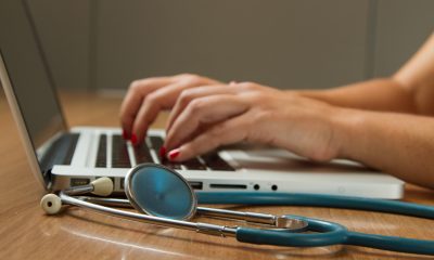 A pair of hands typing on a laptop keyboard. A stephoscope lies next to the keyboard