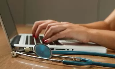 A pair of hands typing on a laptop keyboard. A stephoscope lies next to the keyboard