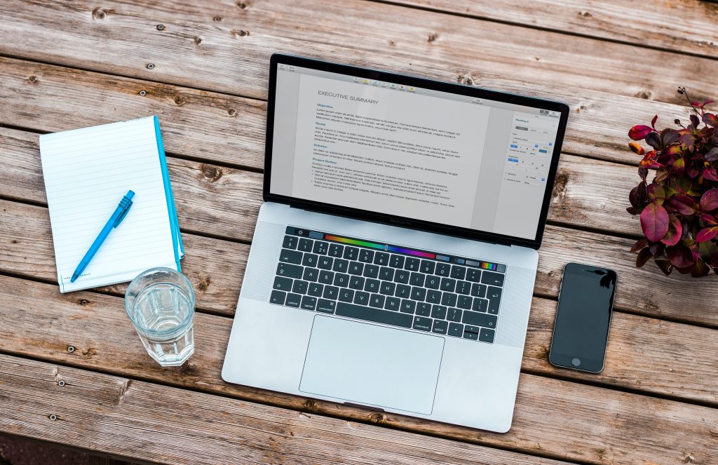 A notepad next to a laptop computer focused on funding. There is a glass of water and phone beside the laptop and purple flowers to the right.