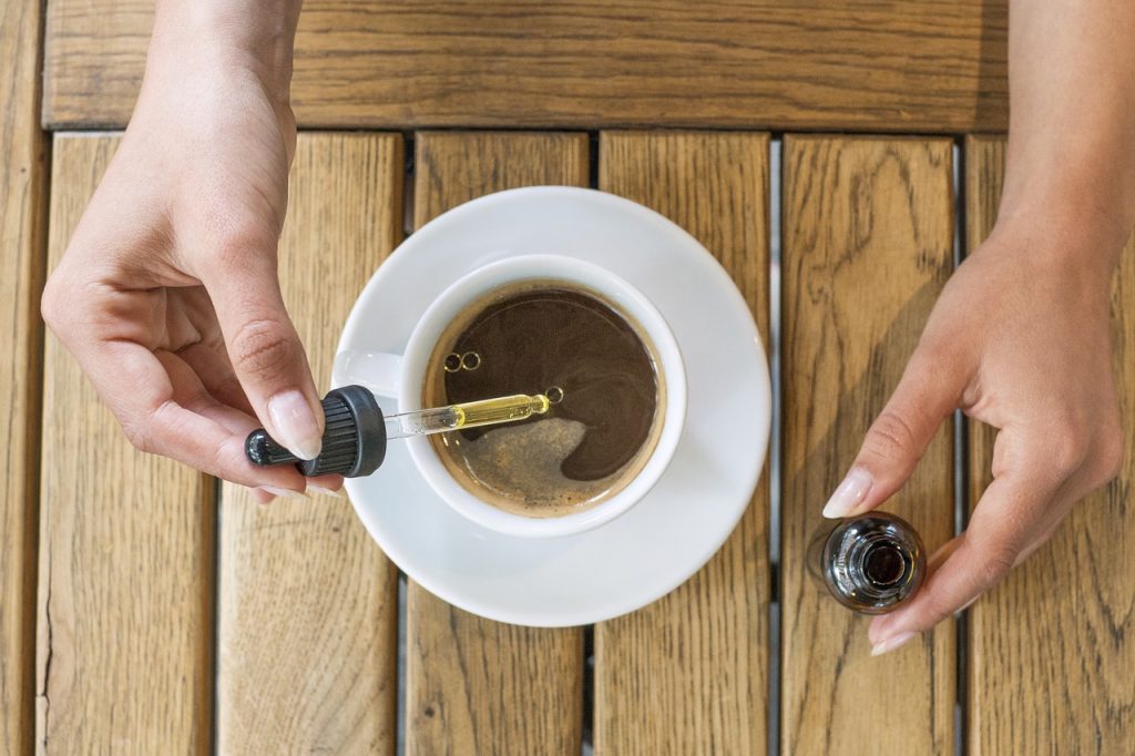 Two hands leaning over a wooden table to put a cbd oil into a drink. There is a cup of coffee in a white cup in the centre of the table.