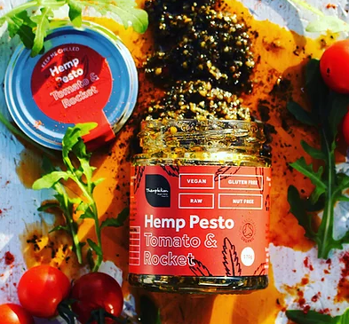A red jar of hemp pesto lies on its side with the lid off. It has spilled the contents which are black and yellow. The label says hemp pesto. There are tomatoes in the photo too