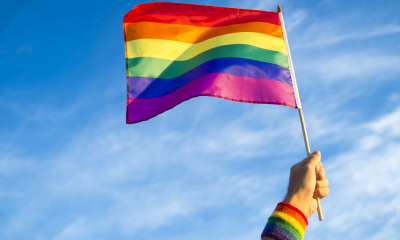 A hand holding a rainbow flag against a blue sky in a celebration of gay pride.