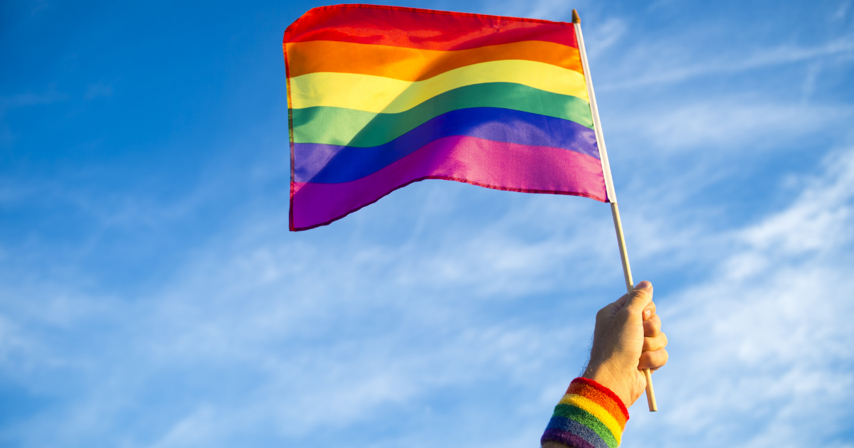 A hand holding a rainbow flag against a blue sky in a celebration of gay pride.