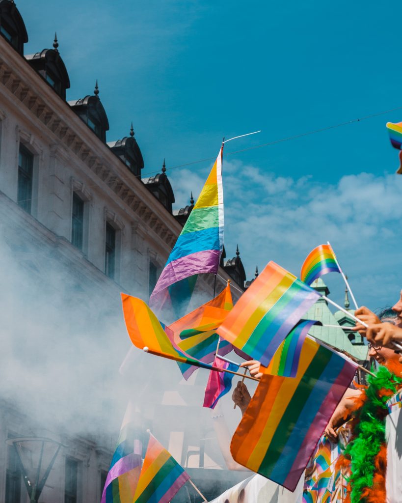 A building against a blue sky. There is a row of brightly coloured gay pride flags and smoke.