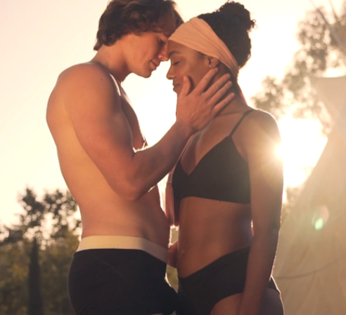 A man in a pair of hemp boxers is about to kiss a girl in a black bra and hemp knickers set. The sun sets in the background