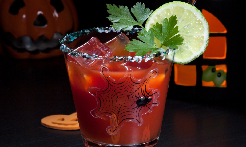 Goblet filled with a Virgin CBD Bloody Mary. Garnished with a lemon slice, basil leaved and a spider. Small lit up haunted house and pumpkin in the background.