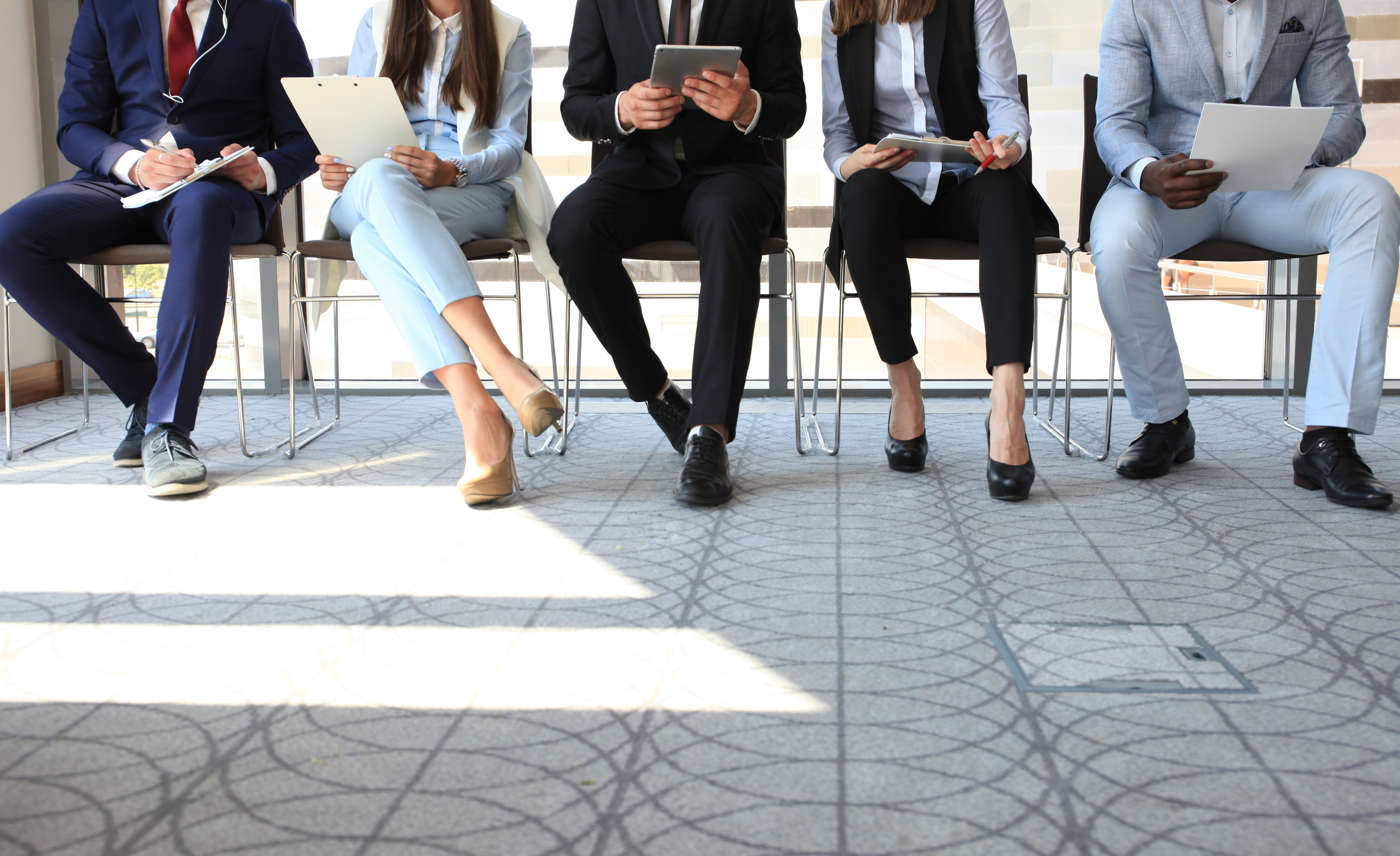 women and men waiting to go into a job interview all sat on chairs using mobile phones